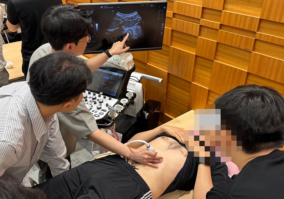 여러 사람이 초음파 기기 주변에 모여 있다. 한 사람이 누워있는 다른 사람의 복부에 초음파 탐촉자를 대고 있고, 또 다른 한 사람이 모니터에 표시된 초음파 영상을 가리키고 있다 — 대한한의영상학회 초음파 약침·도침 강의 | 서울 바로한의원