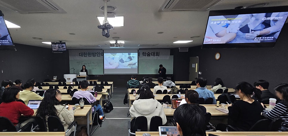 강의실에서 한의학 학술대회 발표자가 강단에 서 있고, 참석자들이 앉아 발표를 듣고 있다. 스크린에는 초음파 유도하 약침 관련 내용이 보인다 — 대한한의영상학회 초음파 약침·도침 강의 | 서울 바로한의원