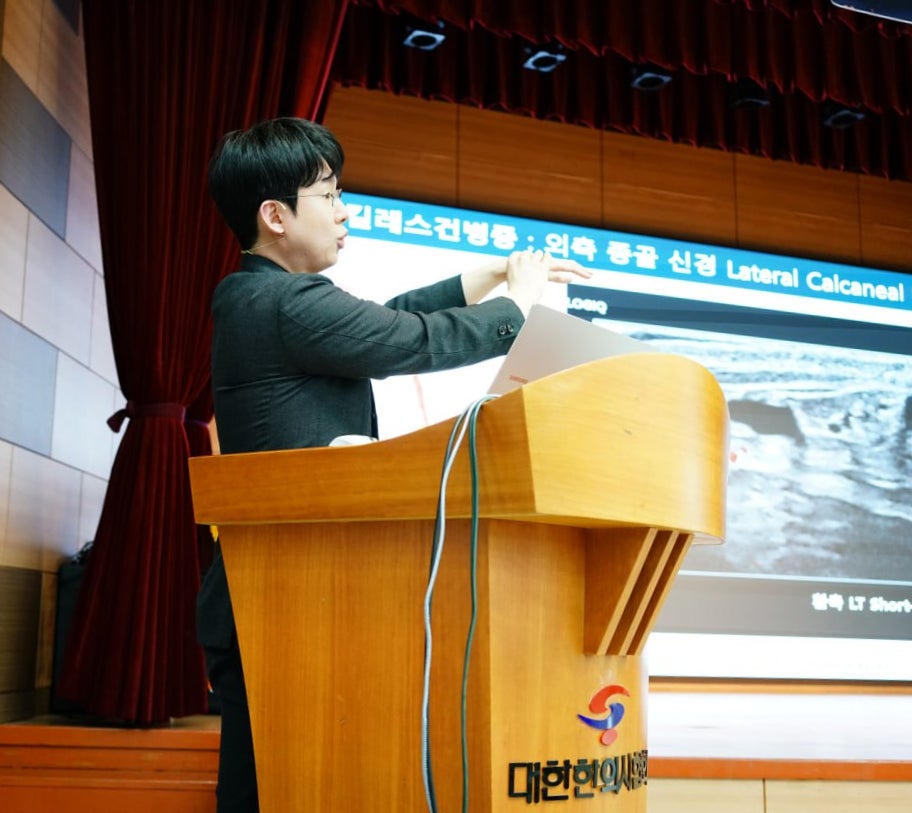 강단에서 발표자가 손으로 제스처를 취하며 설명하고 있고, 뒤쪽 화면에는 아킬레스건병증 관련 내용과 초음파 영상이 보인다. 강단에는 대한한의사협회 로고가 있다 — 대한한의영상학회 초음파 약침·도침 강의 | 서울 바로한의원