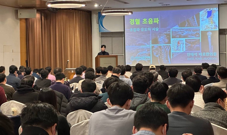 강사가 단상에서 경혈 초음파 관련 내용을 발표하고 있으며, 청중들이 앉아서 강연을 듣고 있는 모습 — 대한한의영상학회 초음파 약침·도침 강의 | 서울 바로한의원