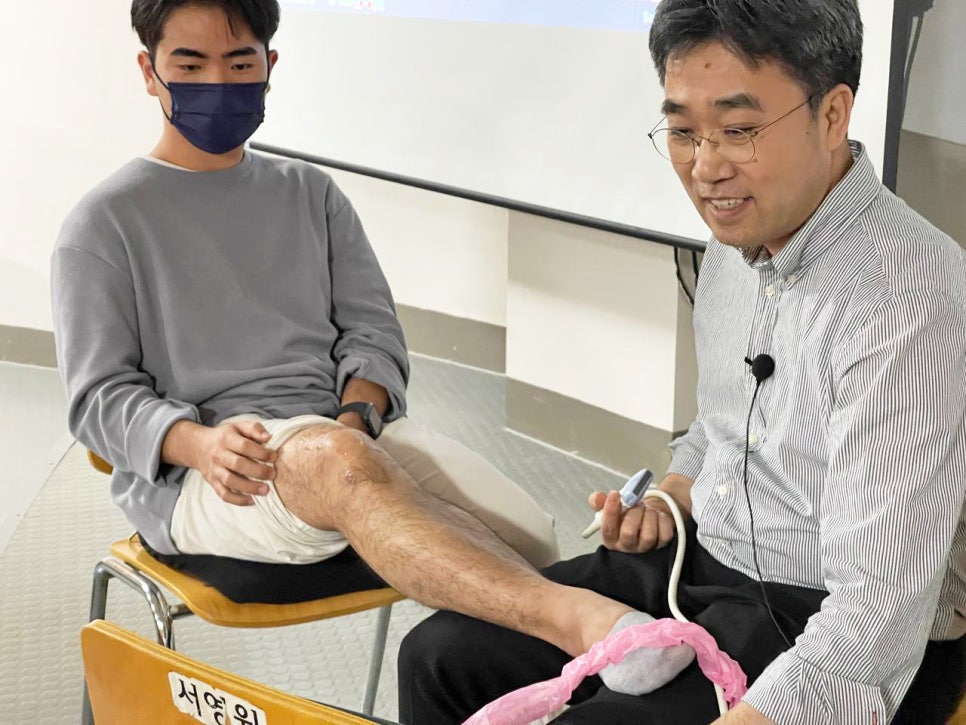 줄무늬 셔츠를 입은 남성이 마스크를 쓴 남성의 다리에 초음파 기기를 대고 시연하는 모습 — 초음파 정밀 진단 | 서울 바로한의원 초음파 약침·도침·침도