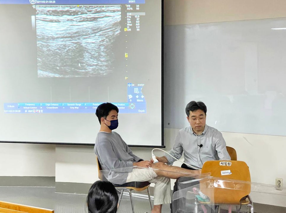 강사가 학생의 다리에 초음파 기기를 사용하여 시연하고 있으며, 스크린에 초음파 영상이 나타나 있다 — 대한한의영상학회 초음파 약침·도침 강의 | 서울 바로한의원