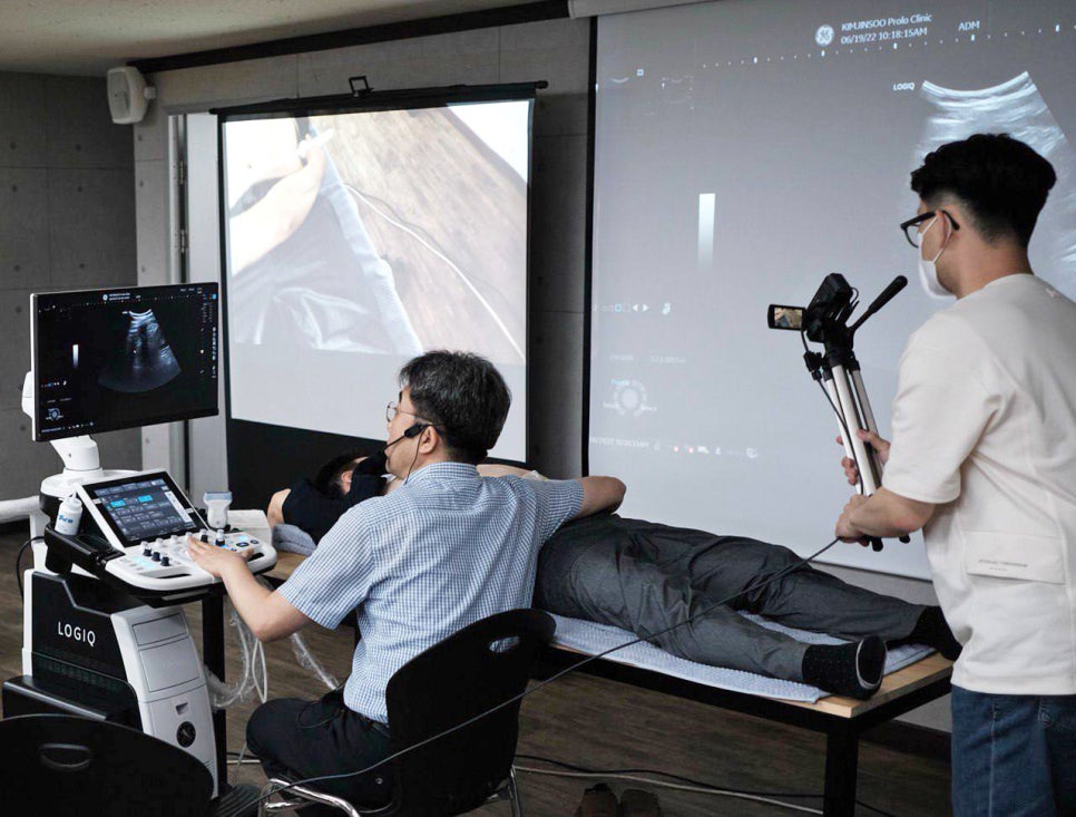 한의사가 엎드린 환자에게 초음파 검사를 하고 있으며, 초음파 영상이 모니터와 스크린에 표시되고 있다. 한 사람이 이 과정을 촬영하고 있다 — 초음파 정밀 진단 | 서울 바로한의원 초음파 약침·도침·침도