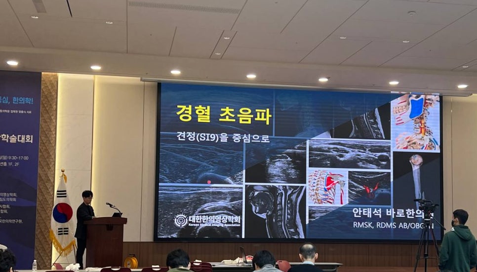 한의학 학술대회에서 강사가 경혈 초음파를 주제로 발표하고 있으며, 스크린에 견정(SI9) 부위의 초음파 영상과 해부학적 그림이 나타나 있습니다 — 대한한의영상학회 초음파 약침·도침 강의 | 서울 바로한의원