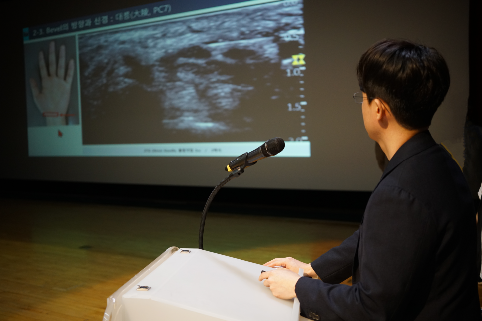 강연자가 마이크가 있는 단상에 서서 손 그림과 초음파 영상이 보이는 스크린을 보고 있다 — 대한한의영상학회 초음파 약침·도침 강의 | 서울 바로한의원