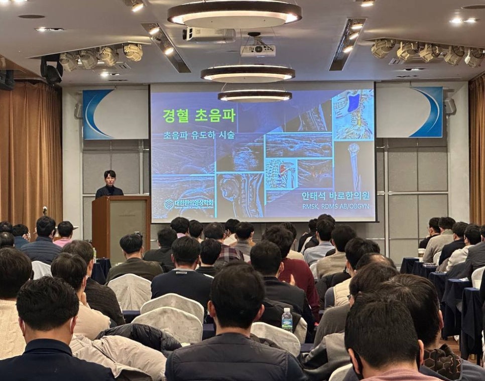 강사가 청중 앞에서 강연하고 있으며, 스크린에는 경혈 초음파와 초음파 유도 시술 관련 내용과 여러 초음파 영상이 보인다 — 대한한의영상학회 초음파 약침·도침 강의 | 서울 바로한의원