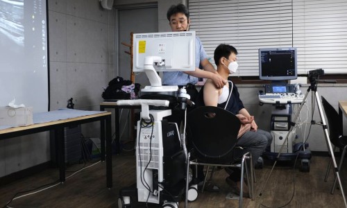 강사가 앉아있는 남성의 어깨에 초음파 기기를 대고 시연하며, 옆 모니터에 초음파 영상이 보인다 — 초음파 정밀 진단 | 서울 바로한의원 초음파 약침·도침·침도