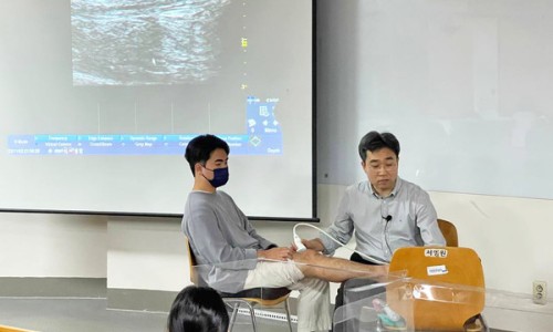 강사가 한 사람의 다리에 초음파 기기를 사용하여 시연하고 있으며, 뒤쪽 화면에 초음파 영상이 보인다 — 대한한의영상학회 초음파 약침·도침 강의 | 서울 바로한의원