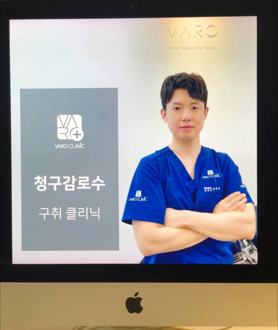 파란색 스크럽복을 입은 남성 의사가 팔짱을 끼고 모니터 앞에 서 있다. 모니터에는 'VARO CLINIC' 로고와 '청구감로수', '구취 클리닉' 문구가 보인다 — 서울 바로한의원 초음파 약침·도침