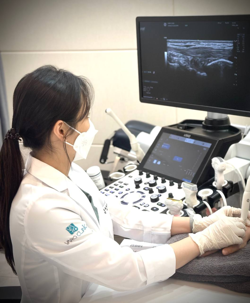 마스크와 장갑을 착용한 의료진이 환자의 손에 초음파 검사를 하고 있으며, 모니터에 초음파 영상이 보인다 — 초음파 정밀 진단 | 서울 바로한의원 초음파 약침·도침·침도