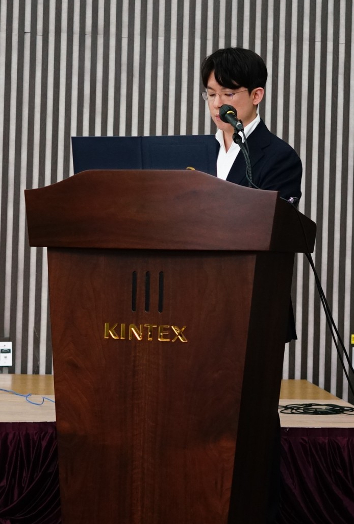 한 남성이 KINTEX 로고가 있는 나무 연단에서 마이크를 잡고 발표하고 있다 — 대한한의영상학회 초음파 약침·도침 강의 | 서울 바로한의원 안태석 원장