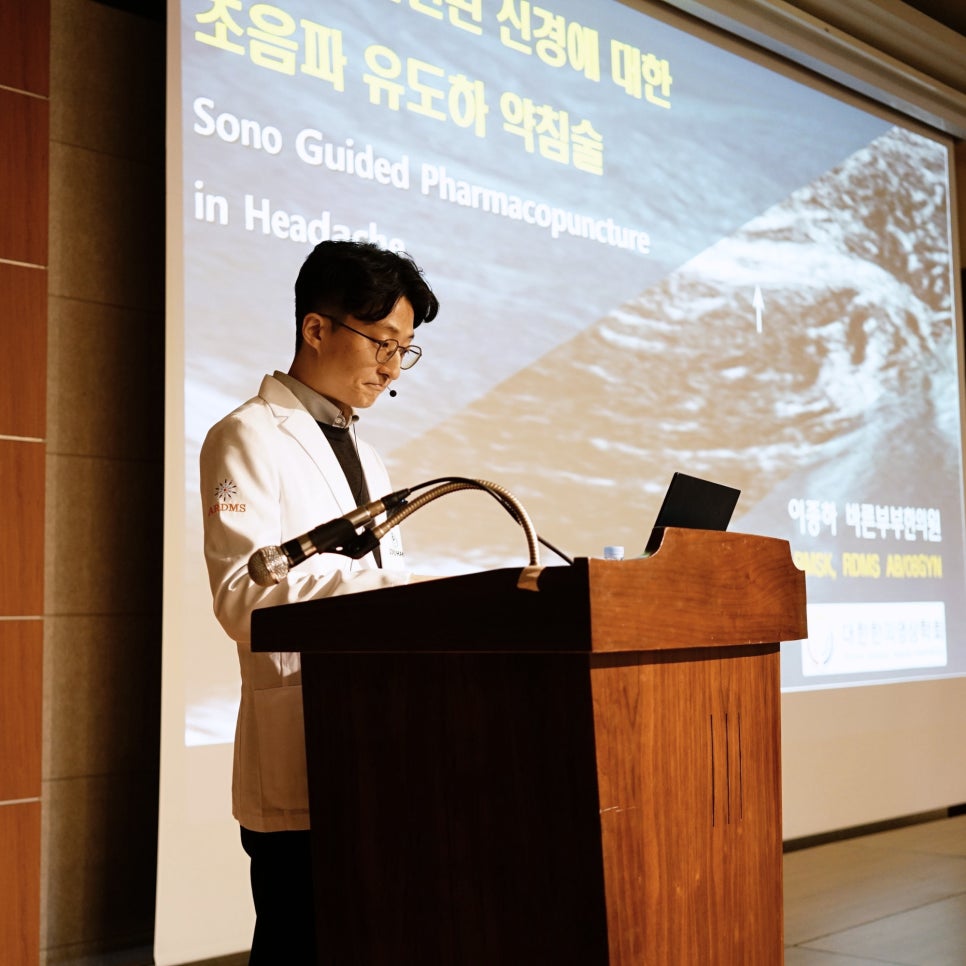 흰색 의사 가운을 입은 남성이 강단에서 발표하고 있으며, 뒤 스크린에는 초음파 유도하 약침술 관련 내용과 초음파 이미지가 표시되어 있다 — 대한한의영상학회 초음파 약침·도침 강의 | 서울 바로한의원 안태석 원장