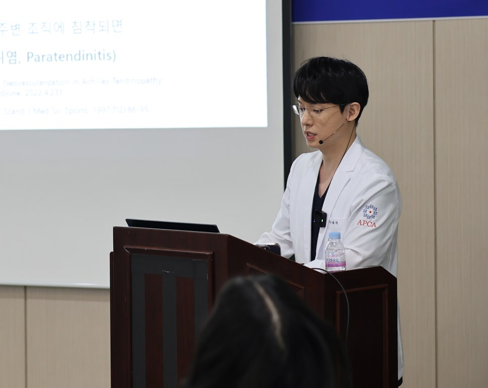 흰색 가운을 입은 남성이 연단에서 마이크를 착용하고 발표하고 있으며, 뒤편에는 글자가 보이는 스크린이 있다 — 대한한의영상학회 초음파 약침·도침 강의 | 서울 바로한의원 안태석 원장