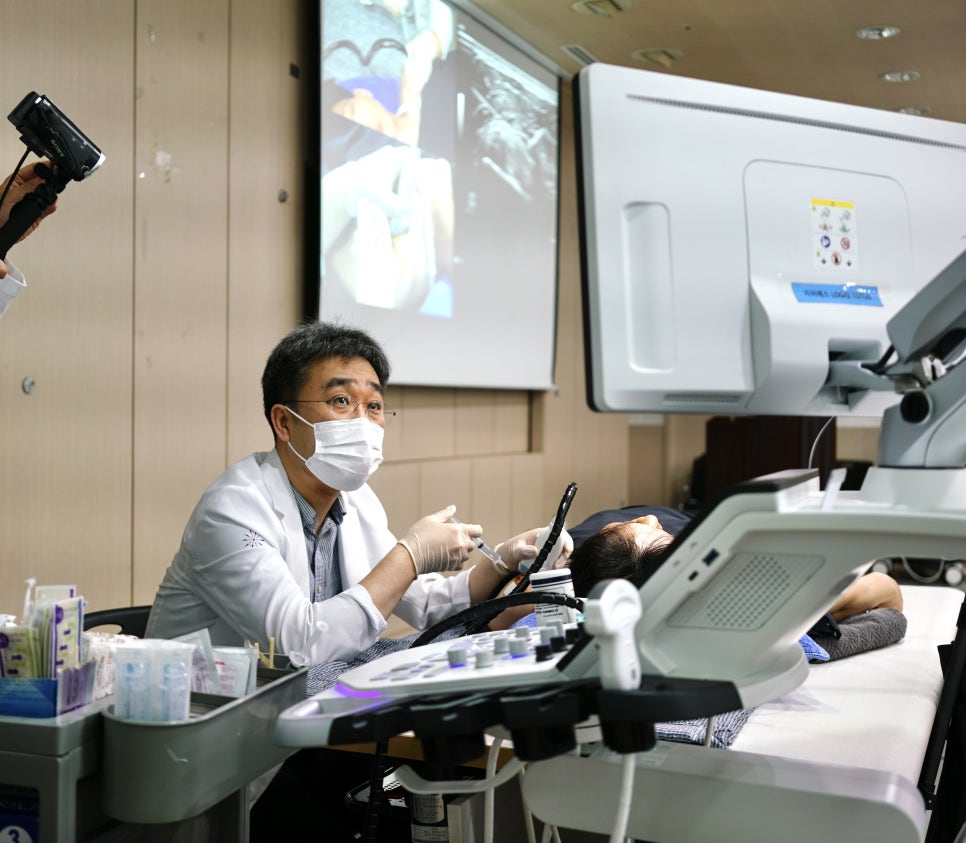 마스크를 쓴 남성 의사가 초음파 기기를 보며 환자에게 시술하고 있다 — 초음파 유도하 약침 시술 | 서울 바로한의원