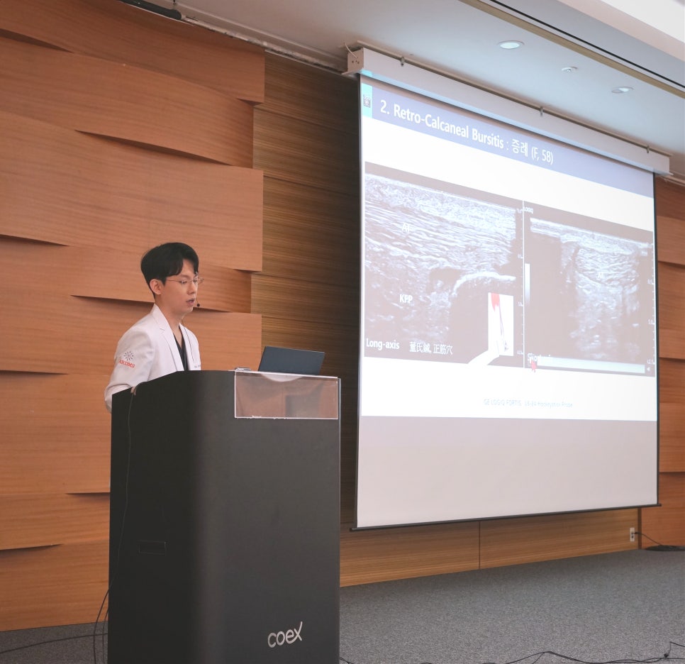 흰색 가운을 입은 남성이 연단에서 발표하고 있으며, 뒤 스크린에는 발목 초음파 영상이 보인다 — 대한한의영상학회 초음파 약침·도침 강의 | 서울 바로한의원 안태석 원장