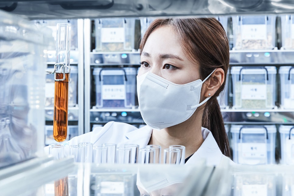 하얀 가운과 마스크를 착용한 여성이 갈색 액체가 담긴 시험관을 들고 있고, 뒤편에는 약재가 담긴 투명 용기들이 선반에 놓여 있다 — 서울 바로한의원 초음파 약침·도침