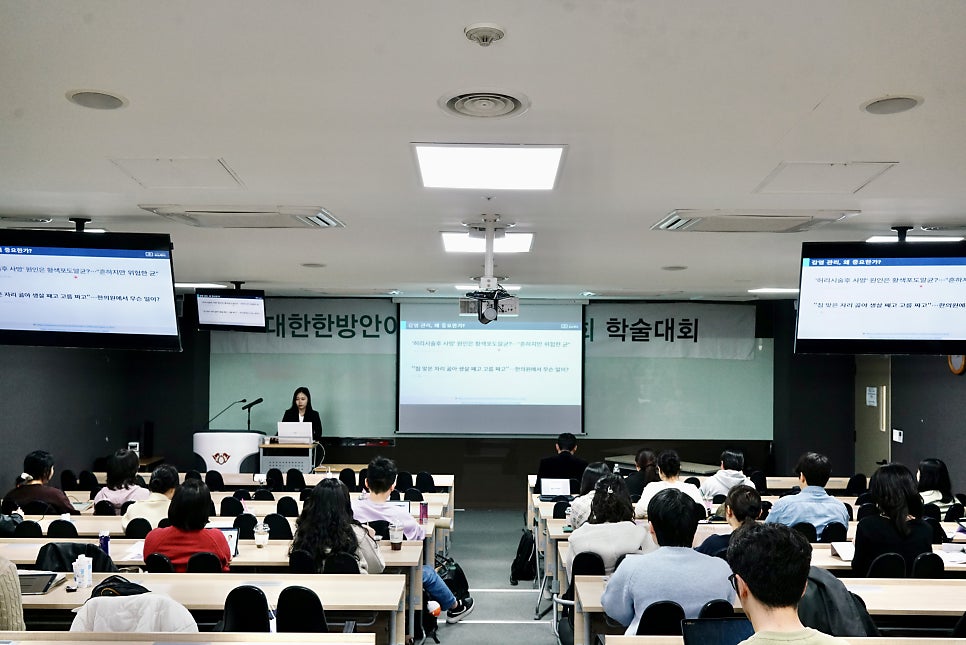 강당에서 한 여성이 발표하고 있으며, 여러 명의 참석자들이 앉아 경청하고 있다 — 대한한의영상학회 초음파 약침·도침 강의 | 서울 바로한의원 문지현 원장