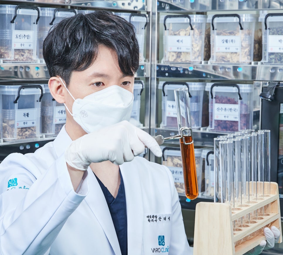 마스크와 장갑을 착용한 한의사가 흰색 가운을 입고 시험관에 담긴 주황색 액체를 집게로 들고 살펴보며, 뒤편에는 한약재가 담긴 용기들이 진열되어 있다 — 초음파 약침·도침 임상 논문 | 서울 바로한의원