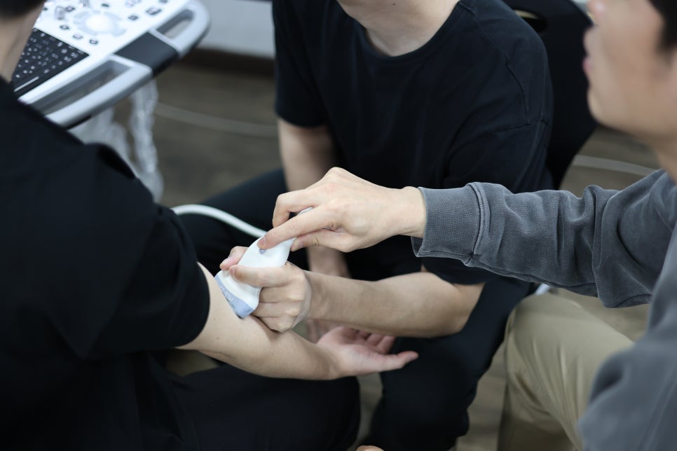 두 사람이 한 사람의 팔에 초음파 탐촉자를 대고 실습하는 모습 — 초음파 유도하 약침 시술 | 서울 바로한의원