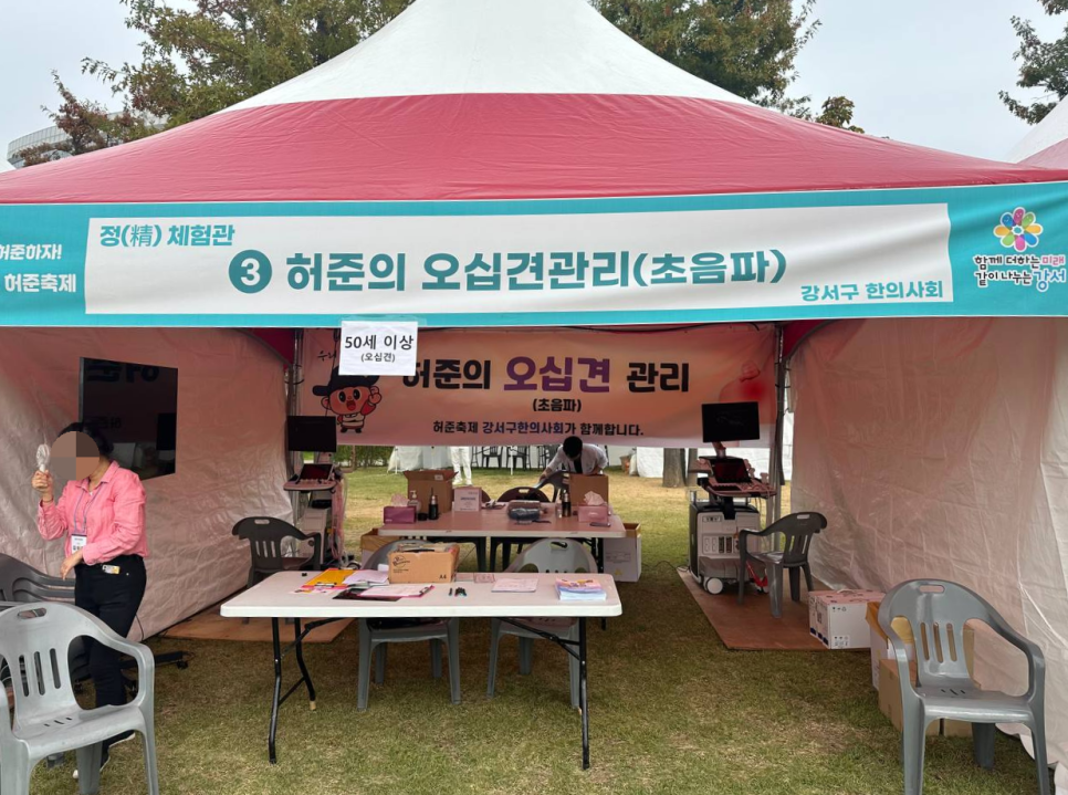 허준축제에 설치된 빨간색과 흰색 천막 부스 내부 모습으로, 테이블과 의자, 초음파 기기가 놓여 있고 한 사람이 서 있다 — 초음파 정밀 진단 | 서울 바로한의원 초음파 약침·도침·침도