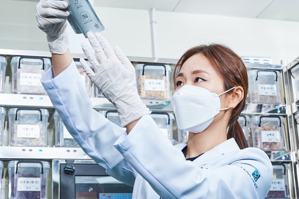 흰색 가운과 마스크를 착용한 사람이 장갑을 낀 손으로 한약 파우치를 들고 약재 선반 앞에서 확인하고 있다 — 서울 바로한의원 초음파 약침·도침