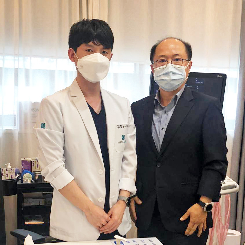 마스크를 착용한 두 남성이 나란히 서 있다. 한 명은 흰색 의사 가운을, 다른 한 명은 정장을 입고 있다 — 서울 바로한의원 초음파 약침·도침