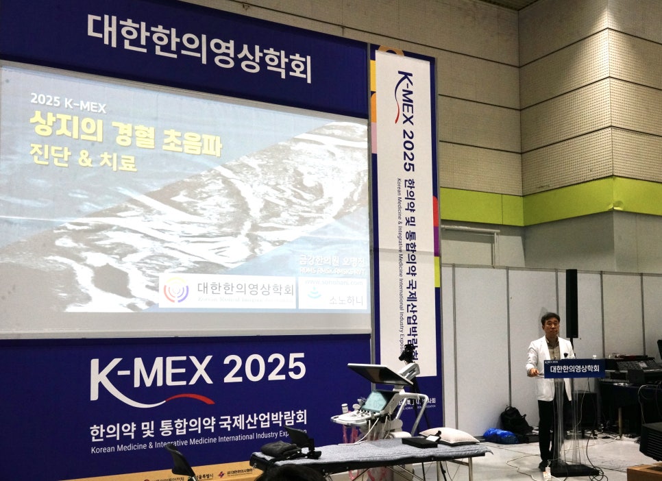 한 남성이 '대한한의영상의학회'와 '상지의 경혈 초음파 진단 & 치료'가 적힌 화면 앞에서 강연하고 있으며, 초음파 기기와 침대가 놓여 있다 — 대한한의영상학회 초음파 약침·도침 강의 | 서울 바로한의원 안태석 원장