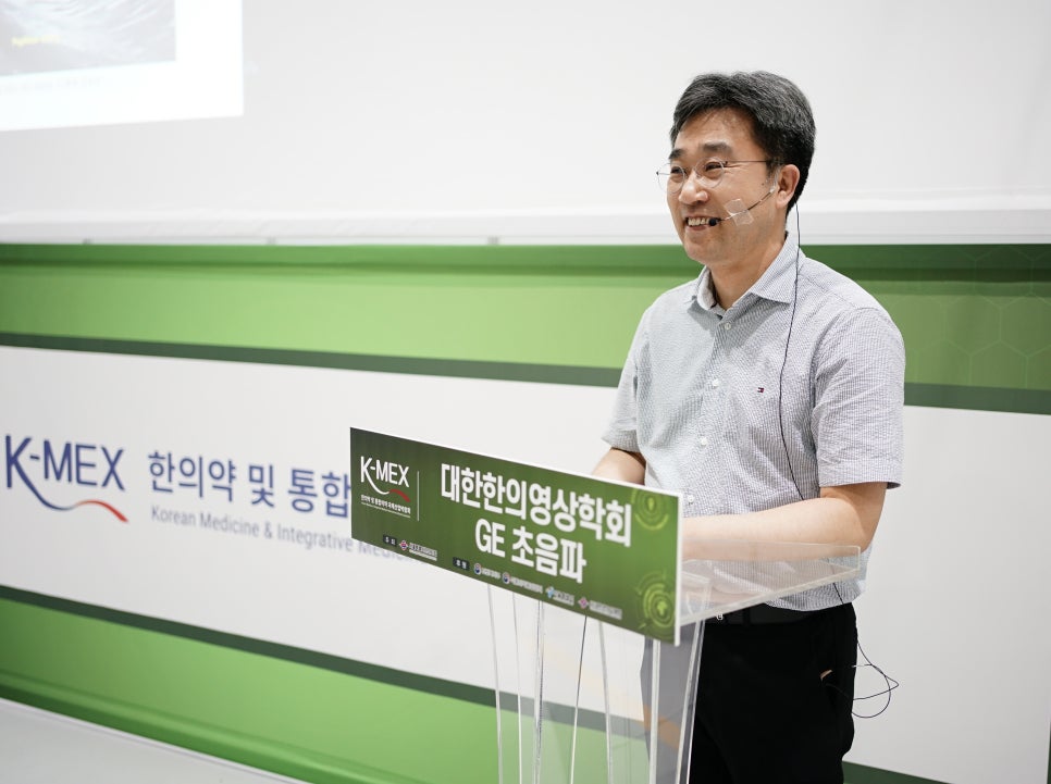 안경을 쓴 남성이 마이크를 착용하고 연단 뒤에서 웃으며 강연하고 있다. 연단에는 '대한한의영상학회 GE 초음파'라고 적힌 녹색 표지판이 있다. 배경에는 'K-MEX 한의학 및 통합' 로고가 보인다 — 대한한의영상학회 초음파 약침·도침 강의 | 서울 바로한의원 안태석 원장