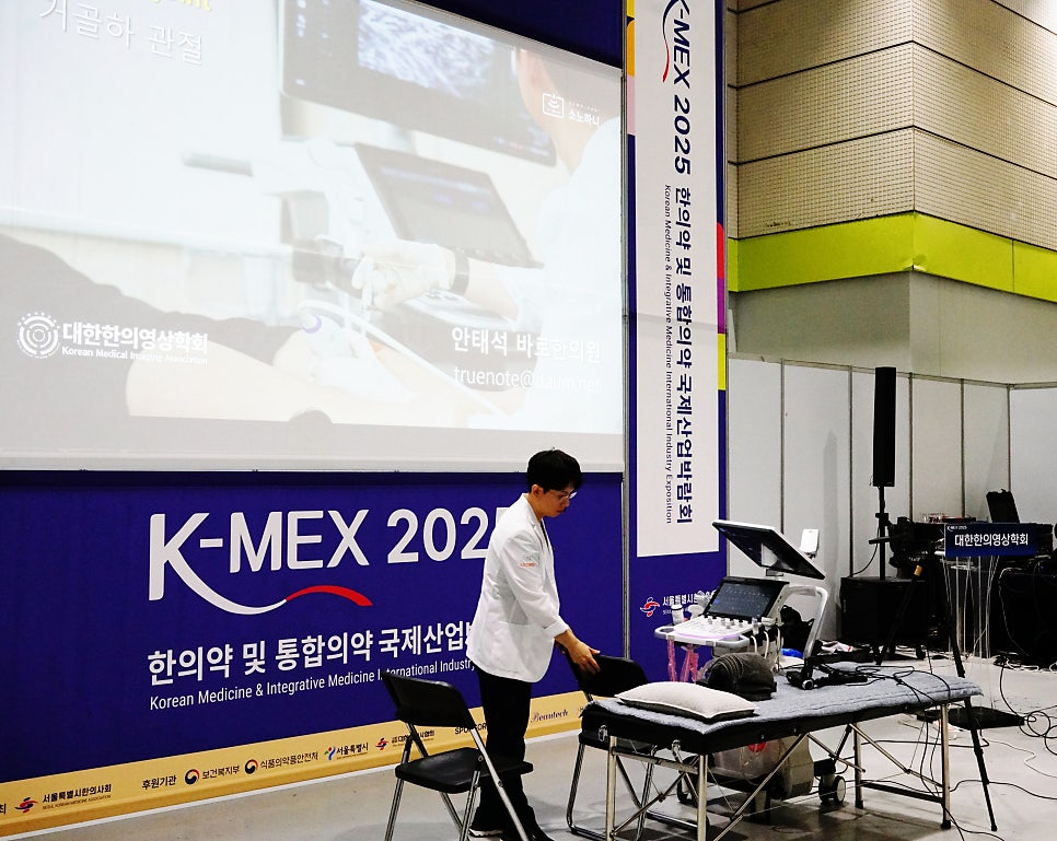 K-MEX 행사장에서 한의사가 초음파 장비와 침대 옆에 서 있고, 스크린에 초음파 관련 영상이 보이는 모습 — 초음파 정밀 진단 | 서울 바로한의원 초음파 약침·도침·침도