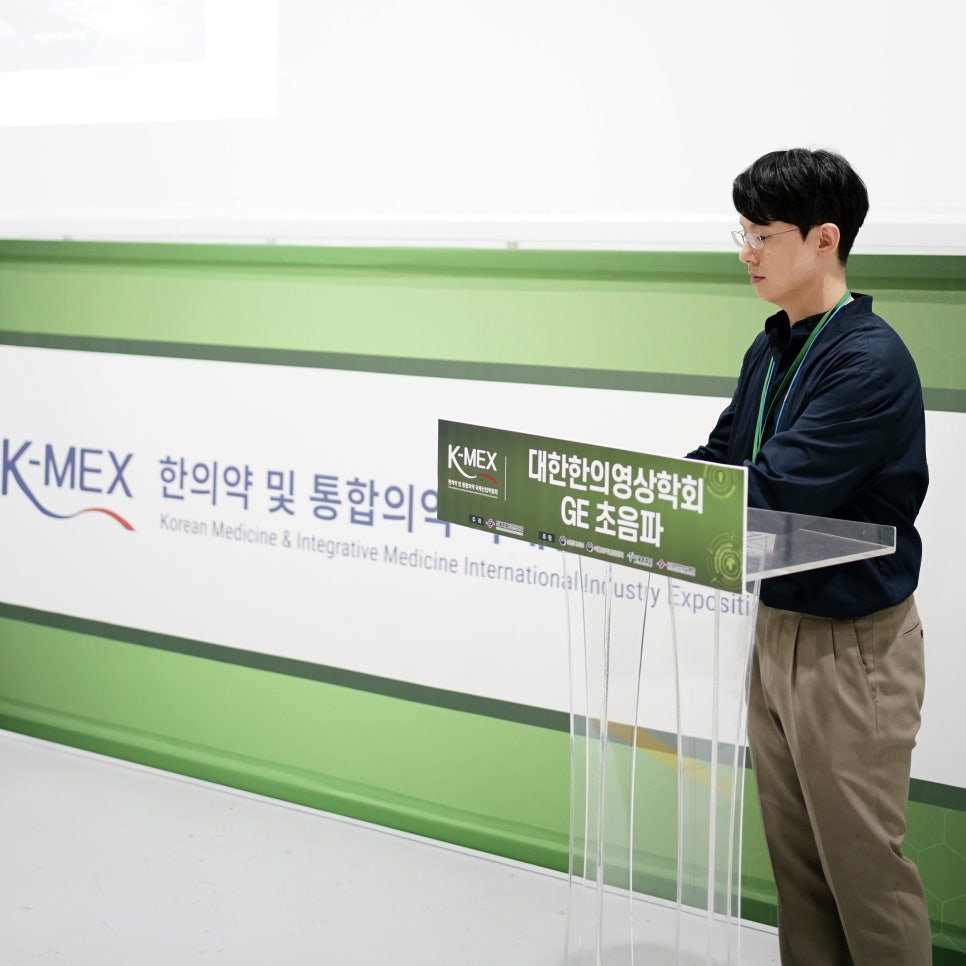 한 남성이 투명한 연단에 서 있고, 연단에는 'K-MEX 대한한의영상학회 GE 초음파'라고 적힌 표지가 놓여 있다. 뒤편에는 'K-MEX 한의약 및 통합의학'이라고 쓰인 현수막이 보인다 — 대한한의영상학회 초음파 약침·도침 강의 | 서울 바로한의원 안태석 원장