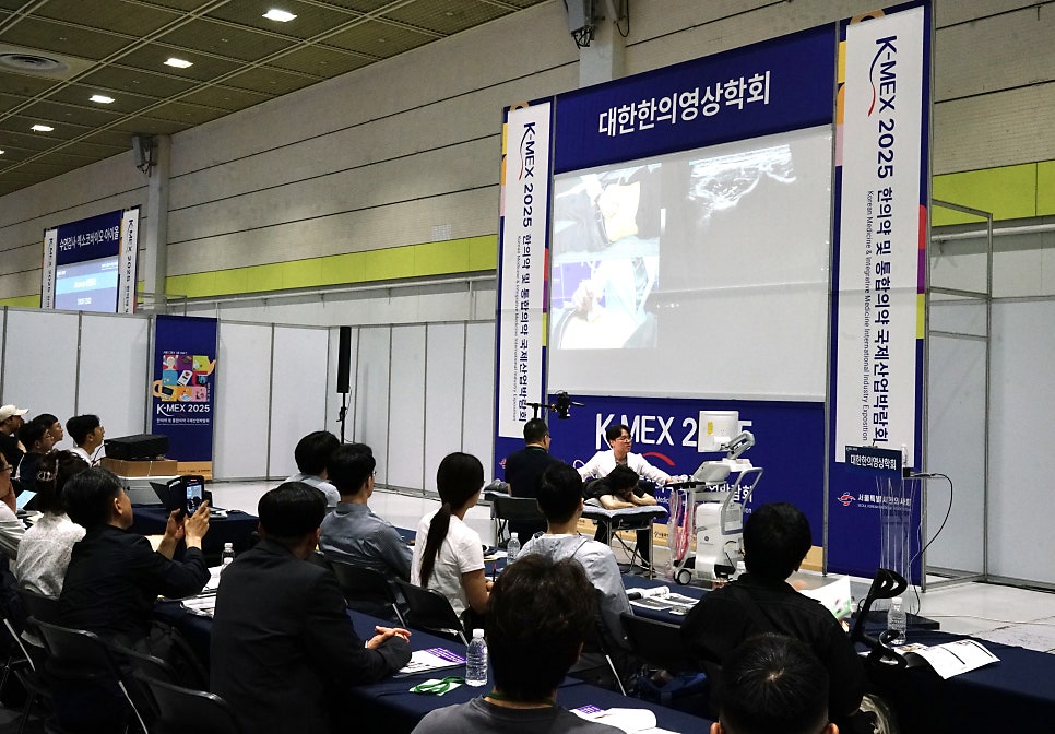 K-MEX 행사에서 강사가 초음파 기기로 시연하는 모습을 청중이 보고 있으며, 대형 스크린에 초음파 영상이 보인다 — 대한한의영상학회 초음파 약침·도침 강의 | 서울 바로한의원