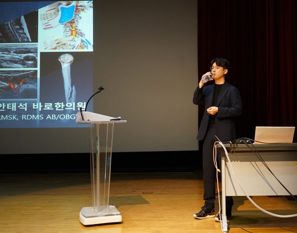 강연 중인 한의사가 물을 마시고 있으며, 뒤 스크린에는 어깨 해부학 및 초음파 이미지가 보인다 — 대한한의영상학회 초음파 약침·도침 강의 | 서울 바로한의원