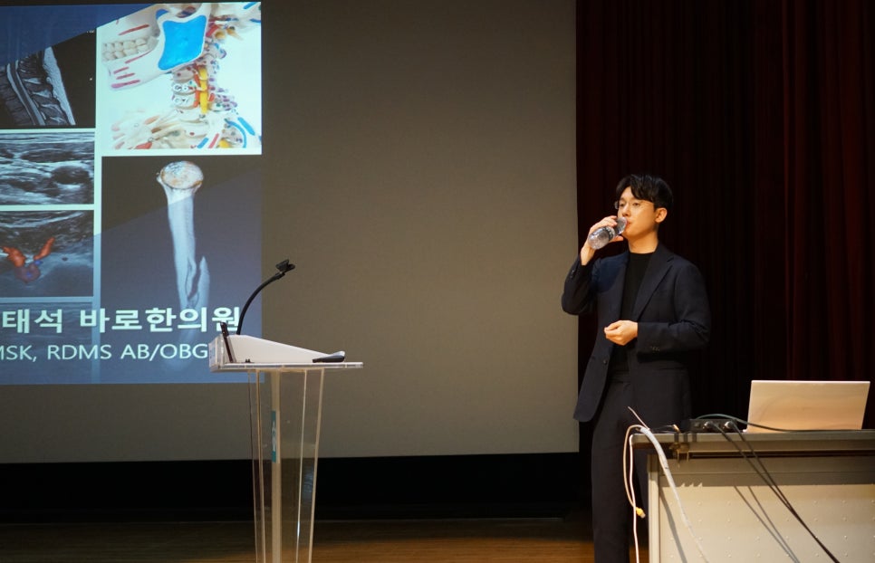 무대 위에서 정장 차림의 남자가 물을 마시고 있고, 뒤편 스크린에는 인체 해부도와 MRI, 초음파 사진 등 다양한 의료 영상이 보인다 — 대한한의영상학회 초음파 약침·도침 강의 | 서울 바로한의원 안태석 원장