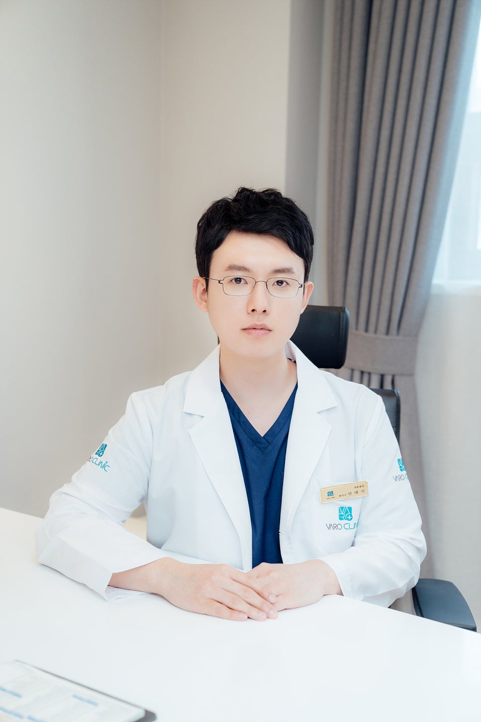 안경을 쓴 남성 한의사가 흰색 의사 가운과 파란색 스크럽을 입고 책상에 앉아 정면을 바라보고 있다 — 서울 바로한의원 초음파 약침·도침