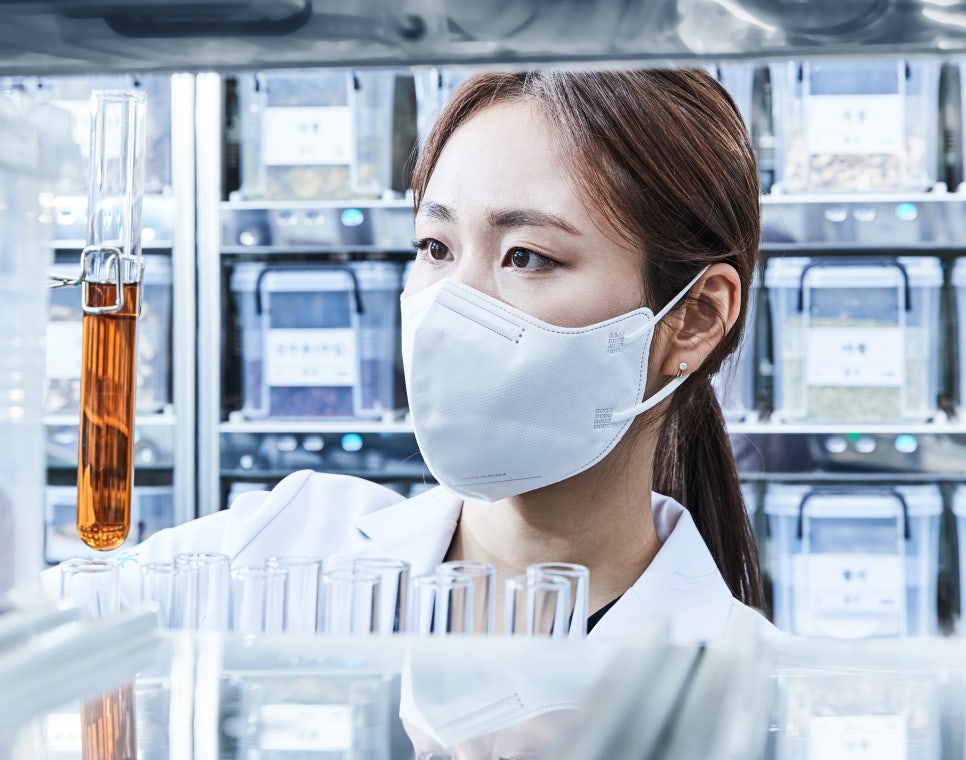 마스크를 착용한 여성이 실험실에서 시험관 속 갈색 액체를 관찰하고 있으며, 뒤편에는 한약재 용기들이 진열되어 있다 — 초음파 약침·도침 임상 논문 | 서울 바로한의원