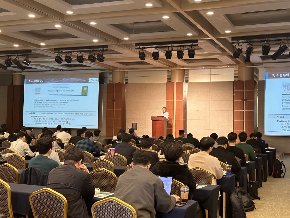 강당에서 강사가 단상에서 발표하고 있으며, 많은 참석자들이 테이블에 앉아 화면을 보고 있다 — 대한한의영상학회 초음파 약침·도침 강의 | 서울 바로한의원