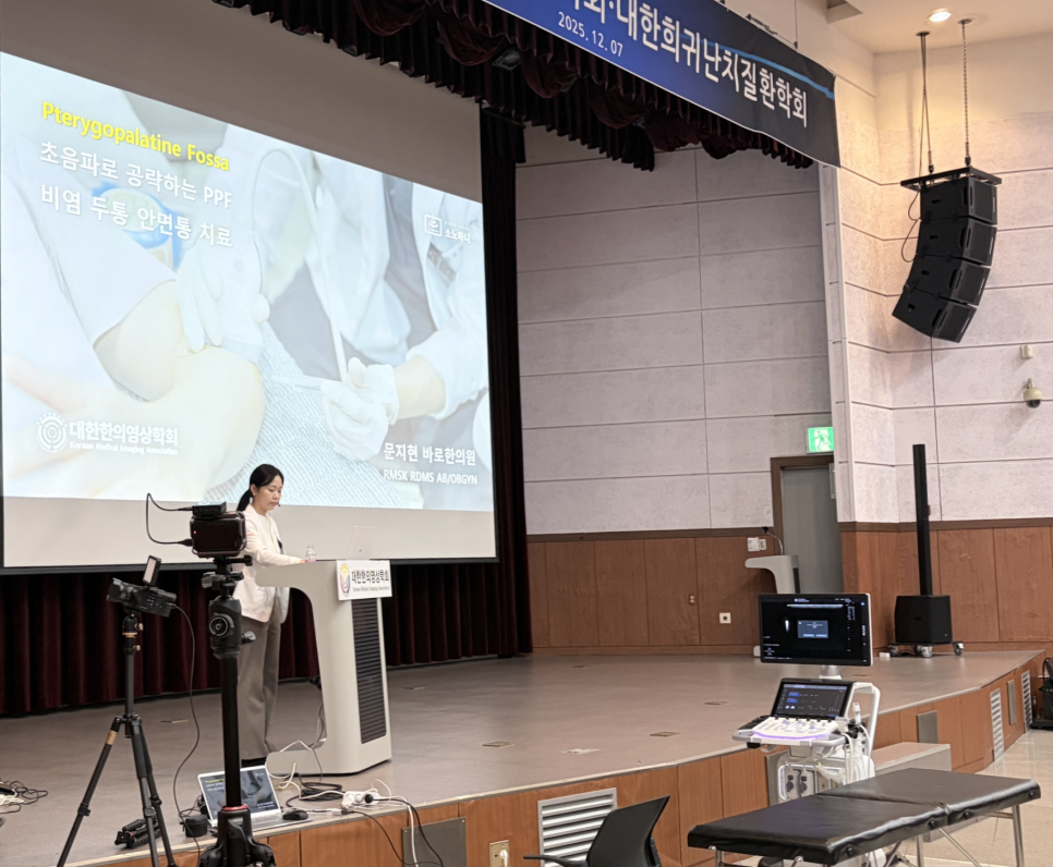 무대에서 강연자가 초음파를 이용한 치료법을 설명하고 있으며, 스크린에는 관련 내용이, 무대에는 초음파 기기와 진료대가 놓여 있다 — 대한한의영상학회 초음파 약침·도침 강의 | 서울 바로한의원