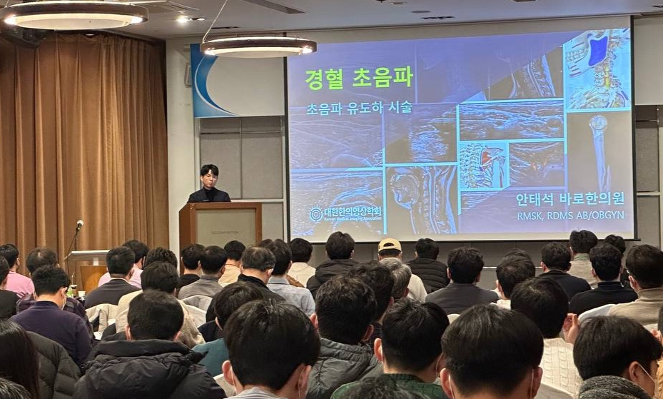 강당에서 연사가 청중에게 경혈 초음파 및 초음파 유도 시술에 대해 강연하고 있으며, 스크린에는 초음파 영상이 보인다 — 대한한의영상학회 초음파 약침·도침 강의 | 서울 바로한의원