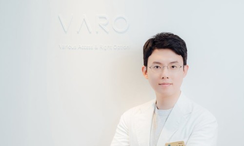 흰색 가운을 입고 안경을 쓴 남성이 'VARO' 로고가 있는 흰색 벽 앞에서 미소 짓고 있다 — 서울 바로한의원 초음파 약침·도침