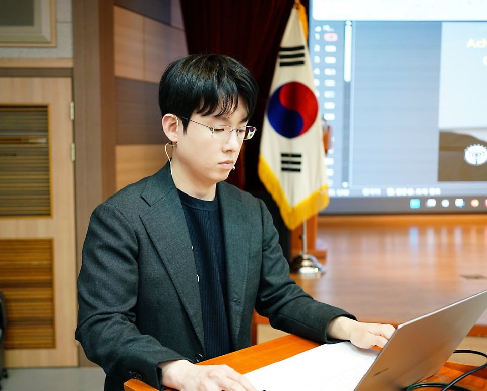 안경을 쓴 남성이 정장 차림으로 노트북을 보고 있으며, 뒤편에 태극기와 화면이 있다 — 대한한의영상학회 초음파 약침·도침 강의 | 서울 바로한의원 안태석 원장