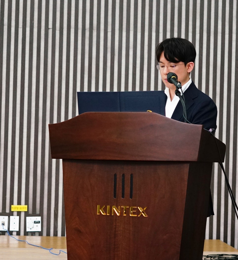 안경을 쓴 남성이 KINTEX 로고가 새겨진 나무 연단에서 마이크를 잡고 발표하는 모습 — 대한한의영상학회 초음파 약침·도침 강의 | 서울 바로한의원 안태석 원장