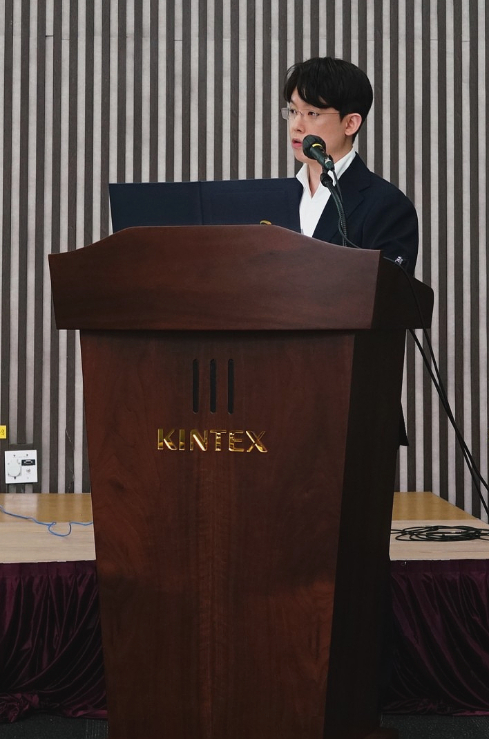 안경을 쓴 남성이 KINTEX 로고가 새겨진 나무 연단에서 마이크를 잡고 말하고 있다 — 대한한의영상학회 초음파 약침·도침 강의 | 서울 바로한의원 안태석 원장
