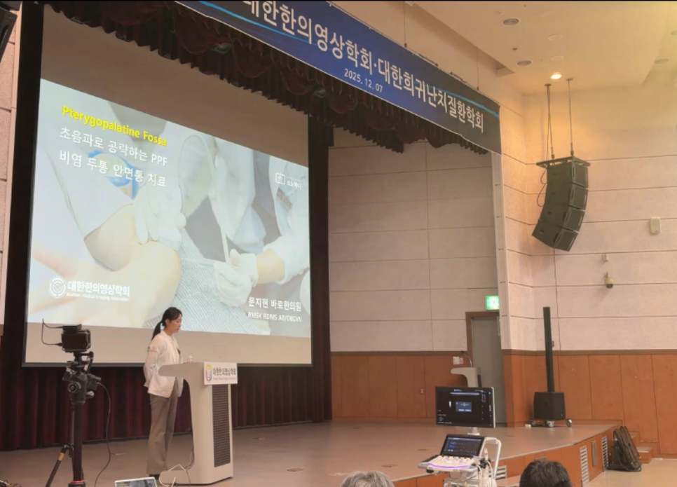 한의학회 강단에서 한 여성이 초음파 가이드 비익구개와 치료 슬라이드를 배경으로 강연하고 있다 — 대한한의영상학회 초음파 약침·도침 강의 | 서울 바로한의원 문지현 원장