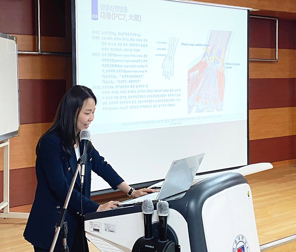 강단에서 여성이 손목 해부학도와 대릉혈(PC7) 정보가 보이는 화면 앞에서 발표하고 있다 — 대한한의영상학회 초음파 약침·도침 강의 | 서울 바로한의원 문지현 원장