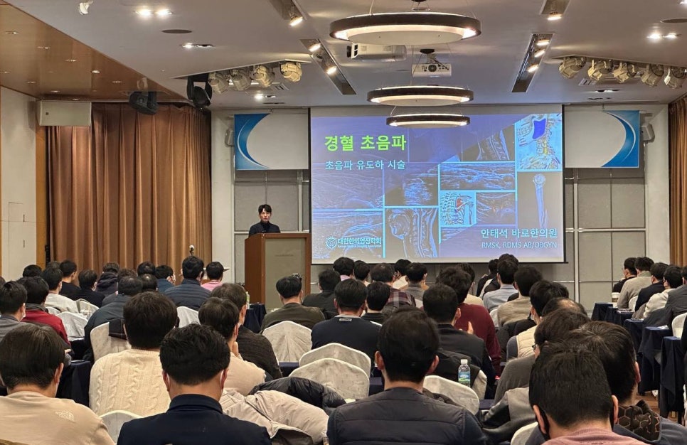 강사가 강연장에서 청중에게 초음파 유도 시술에 대해 발표하고 있다. 스크린에는 '경혈 초음파'와 '초음파 유도하 시술'이라는 문구와 관련 이미지가 보인다 — 대한한의영상학회 초음파 약침·도침 강의 | 서울 바로한의원