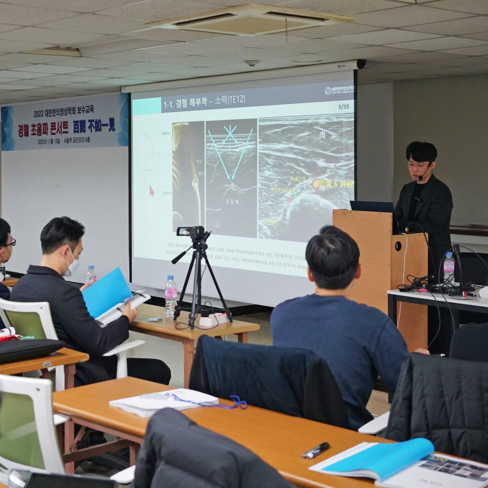 강사가 스크린에 경혈 해부학 및 초음파 이미지를 보여주며 발표하고 있고, 참석자들이 앉아서 보고 있는 세미나 모습 — 대한한의영상학회 초음파 약침·도침 강의 | 서울 바로한의원