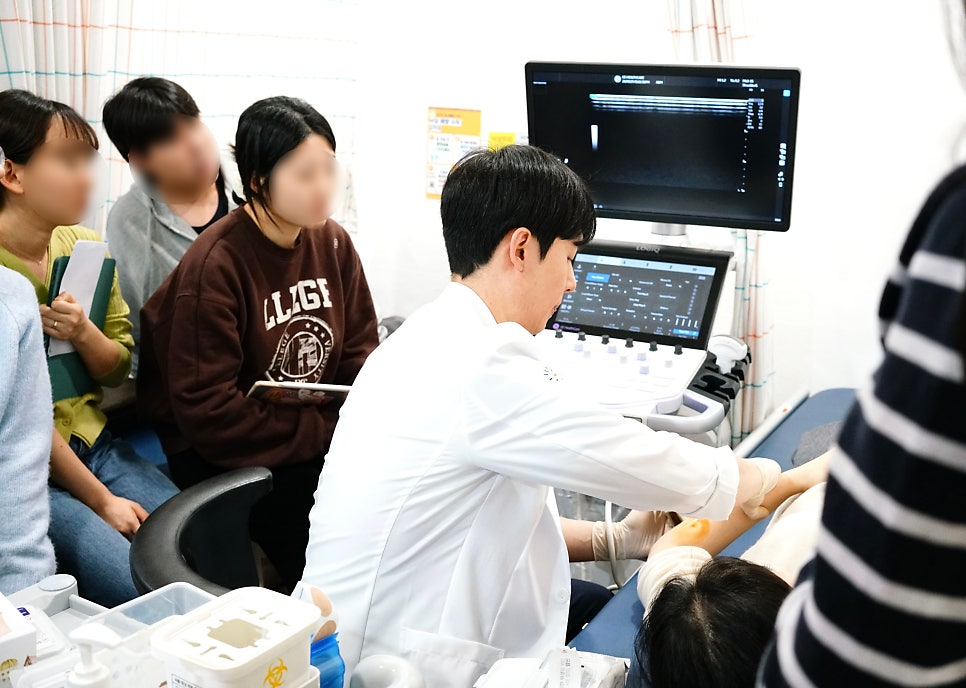 한의사가 환자의 팔에 초음파를 보며 시술하고 있으며, 다른 사람들이 이를 지켜보고 있다 — 초음파 유도하 약침 시술 | 서울 바로한의원