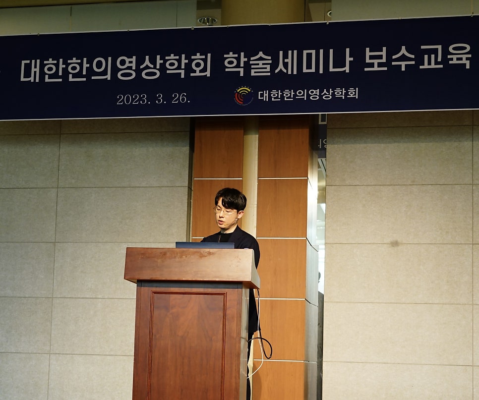 한 남성이 나무 연단에서 발표하고 있으며, 뒤편에는 '대한한의영상학회 학술세미나 보수교육'이라고 적힌 파란색 현수막이 걸려 있다 — 대한한의영상학회 초음파 약침·도침 강의 | 서울 바로한의원 안태석 원장