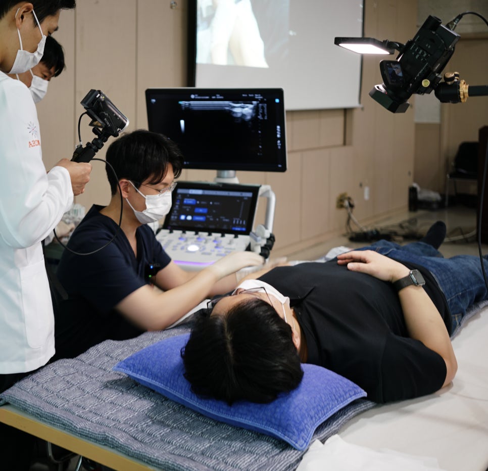 한의사가 환자에게 초음파 기기를 사용하고 있으며, 모니터에 초음파 영상이 보인다. 주변에 다른 사람들이 지켜보고 있다 — 대한한의영상학회 초음파 약침·도침 강의 | 서울 바로한의원