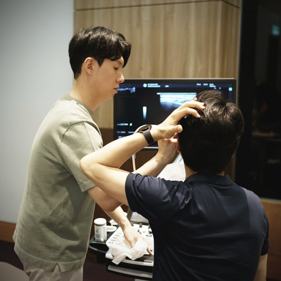 한 남자가 초음파 기기 앞에서 서서 초음파 화면을 보고 있고, 다른 남자는 앉아서 화면을 보고 있다 — 초음파 정밀 진단 | 서울 바로한의원 초음파 약침·도침·침도